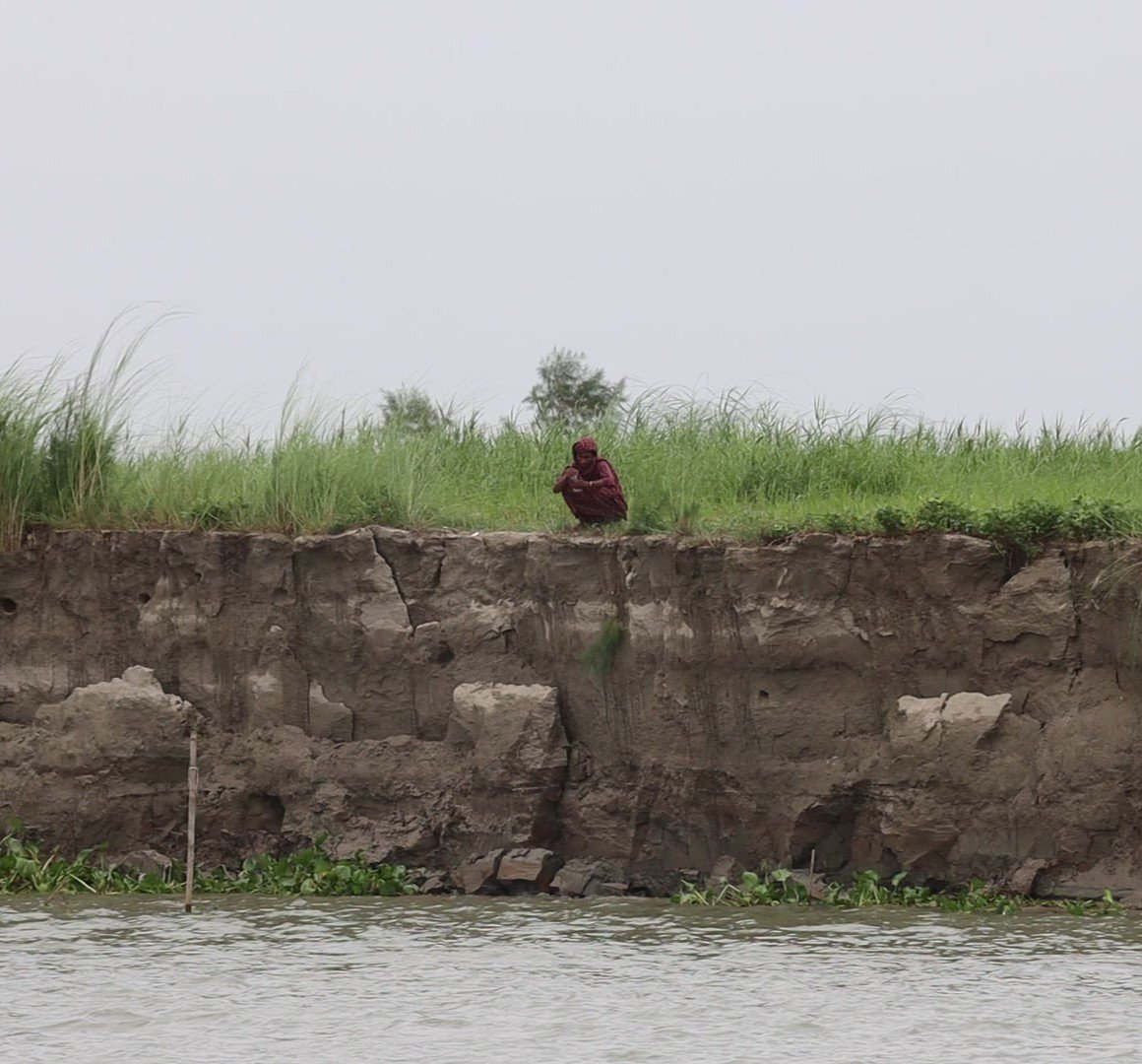 A woman sitting at the grassy edge of a riverbank that has been sheared away by erosion