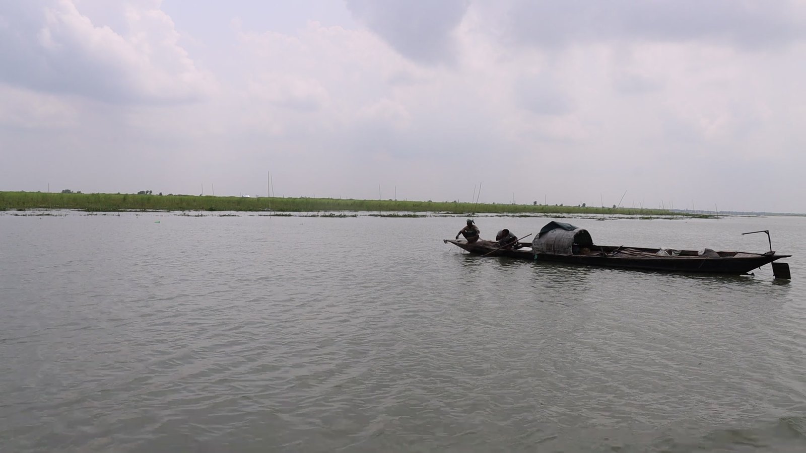 Two fishermen poling a wooden boat across a wide river under a pale monsoon sky